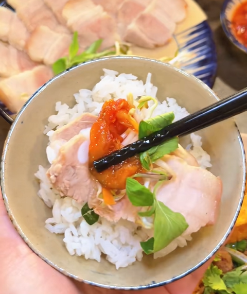 A close-up of a hand holding a bowl of rice topped with sliced meat, red sauce, and greens, with black chopsticks resting on top, alongside a plate of sliced meat.