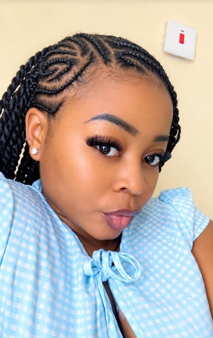 A woman with intricate braided hairstyle and a blue gingham top poses for the camera.