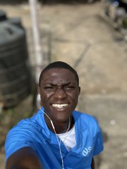 A young man in a blue t-shirt with earphones smiles broadly while taking a selfie outdoors.