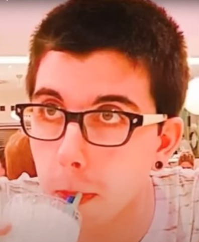 A close-up selfie shows a young person with short dark hair and black-rimmed glasses drinking from a cup with a straw.