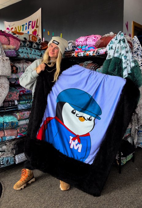 A woman wearing a beanie holds up a blue blanket featuring a penguin character wearing a cap with 'KM' on its outfit, in a shop filled with blankets.