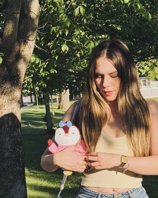 The image features a woman standing next to a tree, holding a stuffed animal that looks like a penguin with a pink bow around its neck. The woman appears to be wearing a yellow tank top and blue jeans. In the background, there is a house visible behind her.