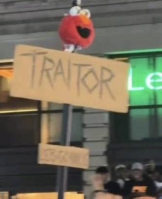 An Elmo puppet sits atop a hand-written cardboard sign reading "TRAITOR" during what appears to be a public demonstration, with a crowd visible below.
