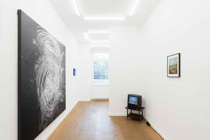 A minimalist white-walled art gallery space featuring a large black abstract painting, a small framed artwork, and an old CRT television on a wooden stand.