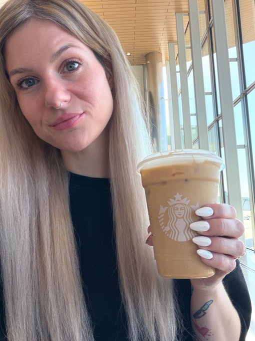 A selfie of a young blonde woman with a nose ring holding an iced coffee in a Starbucks cup, showing her white manicured nails and a small tattoo on her wrist in a bright indoor setting.
