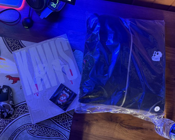 A flattened jacket with a skull patch and a poster featuring the text 'SKATERS' in plastic packaging, laid on a wooden desk in blue ambient lighting.