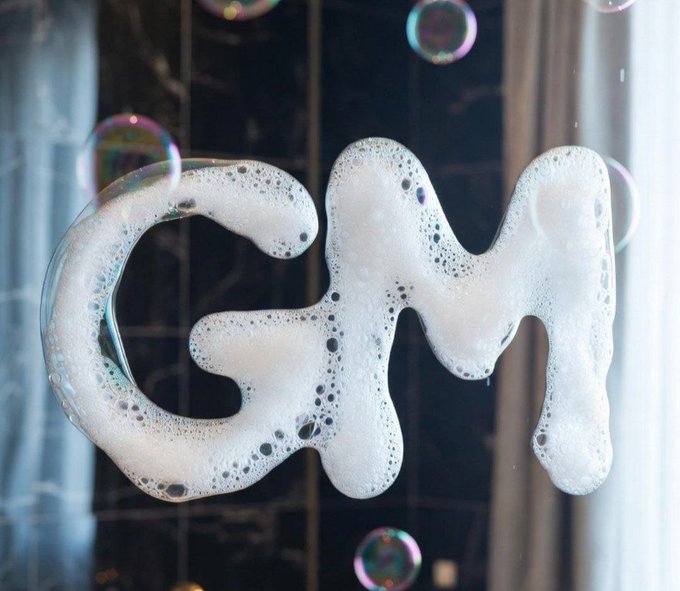 A close-up shot of the word 'GM' written in bubbly white soap suds on a glass surface, accompanied by floating iridescent soap bubbles.
