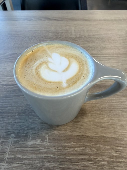 A latte with leaf-shaped foam art sits in a gray ceramic mug on a wooden table.