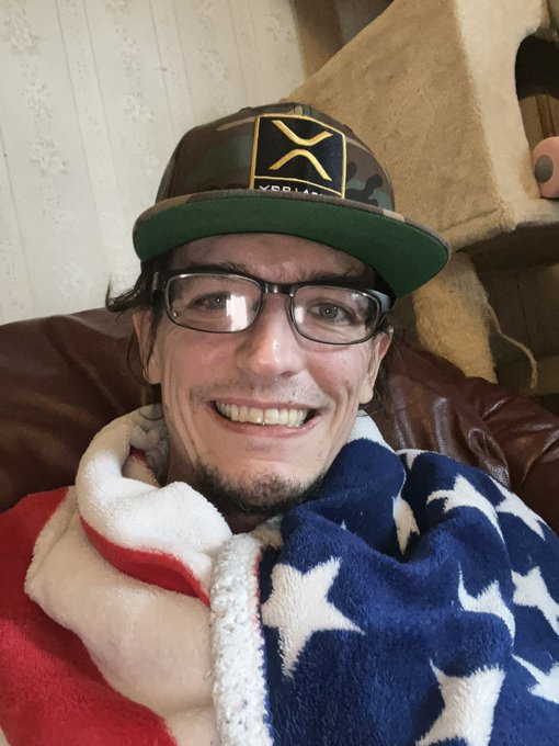 A selfie of a smiling man wearing glasses, a camo and green trucker hat with an X logo, and wrapped in an American flag blanket.