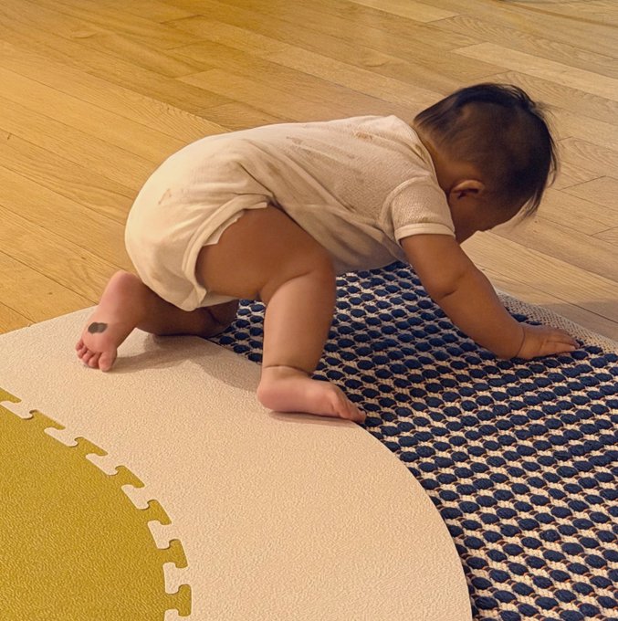 A baby in a white onesie crawls on a textured mat with a blue dotted pattern and yellow edge pieces, placed on a light wooden floor.