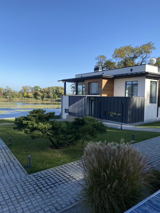 A modern two-story house with black and wood exterior paneling, situated beside a lake with a manicured lawn and pine tree in the foreground.