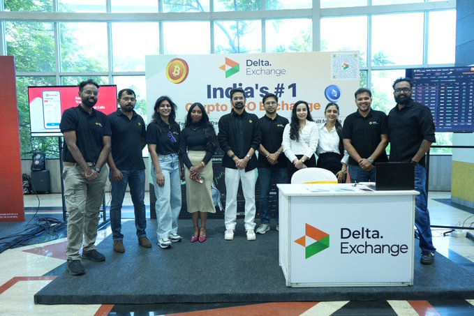 A group of nine people pose for a photo at a Delta Exchange promotional booth, with signage reading 'India's #1 Crypto USD Exchange'.