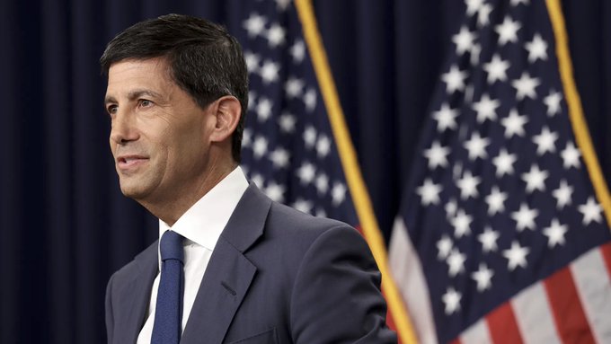 A man in a dark blue suit and tie stands at a podium with American flags in the background.