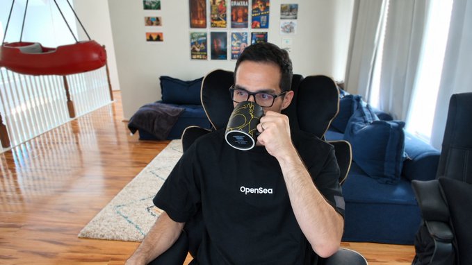 A man wearing glasses and an "OpenSea" t-shirt sips from a dark mug while seated in a gaming chair in a living room with movie posters on the wall. The relaxed, casual pose pairs with the "GM" (good morning) post, conveying a chill start-of-day vibe.