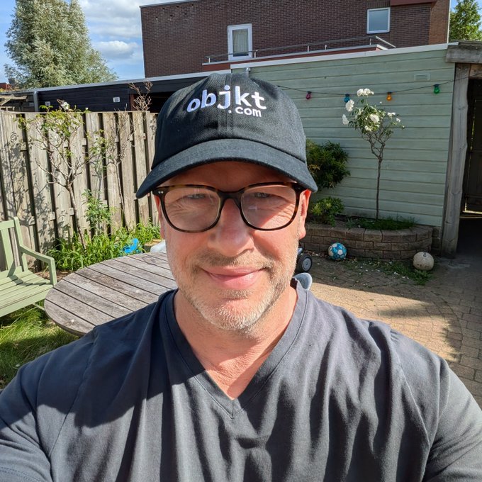 A man in a backyard wearing a new black cap featuring the "objkt.com" logo, matching the post about his new cap from @objktcom.