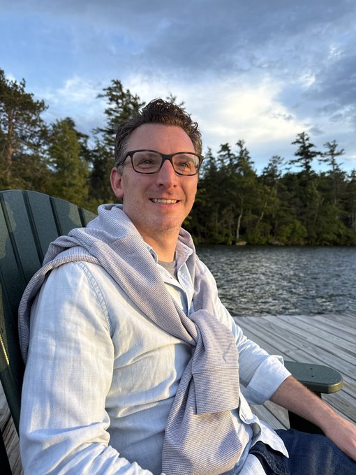 The image features a man sitting in a green chair with his legs crossed, smiling for the camera. He is wearing glasses and has a white shirt on. The setting appears to be outdoors, possibly near a lake or river, as there are trees visible in the background. The caption of the image includes mentions of @BTC_MS_THRIVING, @BeantownBitcoin, and @NEEDcreations, which suggests that this photo might have been taken during an event related to cryptocurrency or simply capturing a memorable moment outdoors with friends.
