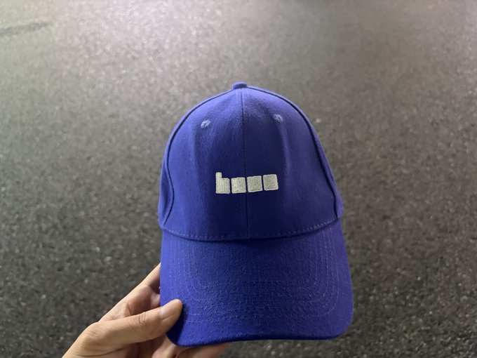 A hand holds a blue baseball cap with a small white logo resembling four squares or rectangles on the front.
