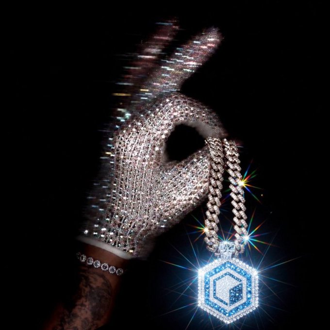 A gloved hand encrusted with sparkling diamonds forms a peace sign while holding a thick diamond chain with a hexagonal pendant against a black background.