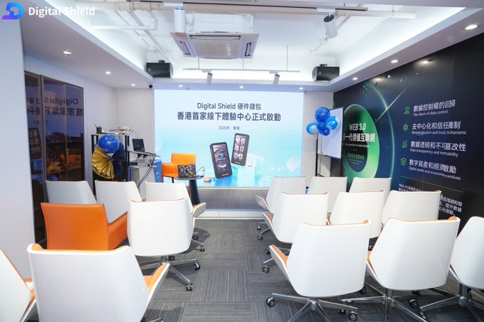 An indoor presentation room setup for 'Digital Shield' event with a screen displaying Chinese text including '香港首家降下體驗中心正式啟動', white and orange chairs arranged for an audience, and promotional balloons.