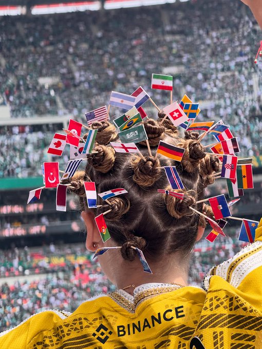 The image shows a person with their hair styled into a bunch of small flags or pins attached to their head. The person's hair appears to be tangled due to the numerous flags. In the background, there are several other people present at an event, possibly attending a sports game or a similar gathering.