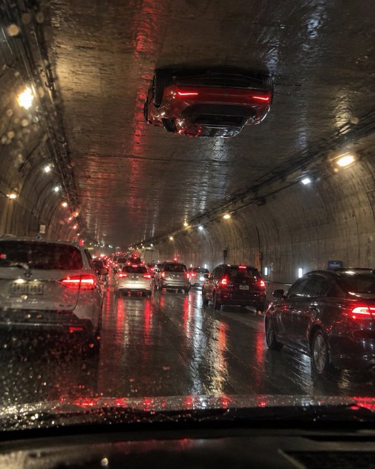 A nighttime view from inside a car driving through a wet tunnel with heavy traffic, featuring an inverted car reflection on the wet ceiling above the windshield.