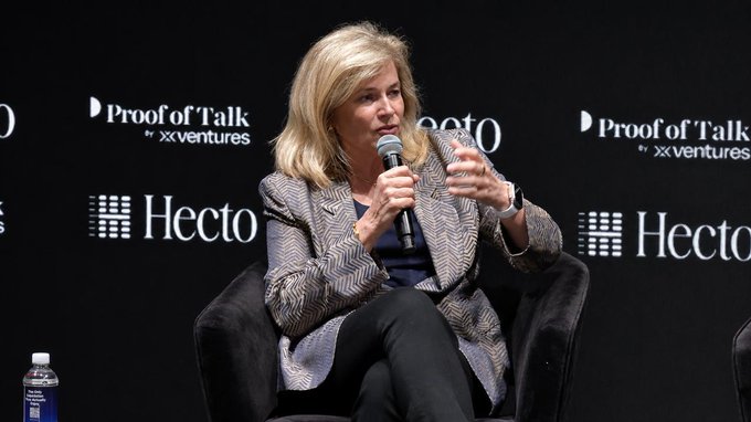 The image features a woman sitting in a chair, holding a microphone and speaking into it. The background of the image includes a black wall with white text that reads "ProfitOfTalks" and "HECTO". There are also two water bottles visible on the floor next to her.