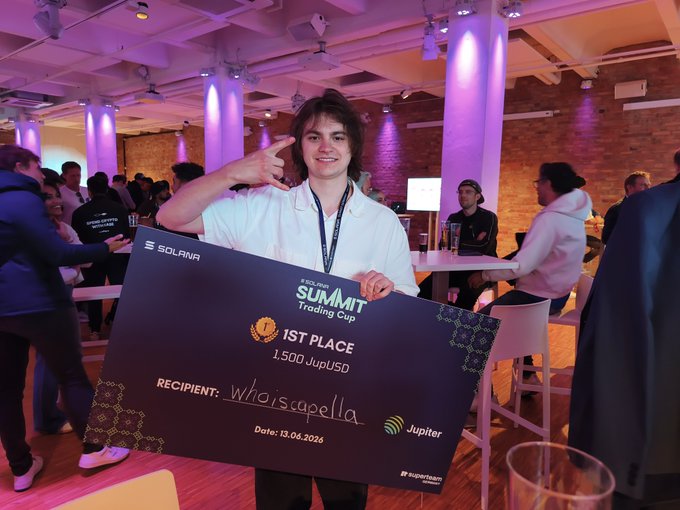 The image features a young man holding up a black card with white text that reads "1st place" and "Winning prize of 500€". The man is standing next to a group of people, some of whom are sitting at tables while others stand around him. There are also several chairs visible in the scene. In addition to the main subject, there's another person partially visible on the right side of the image.