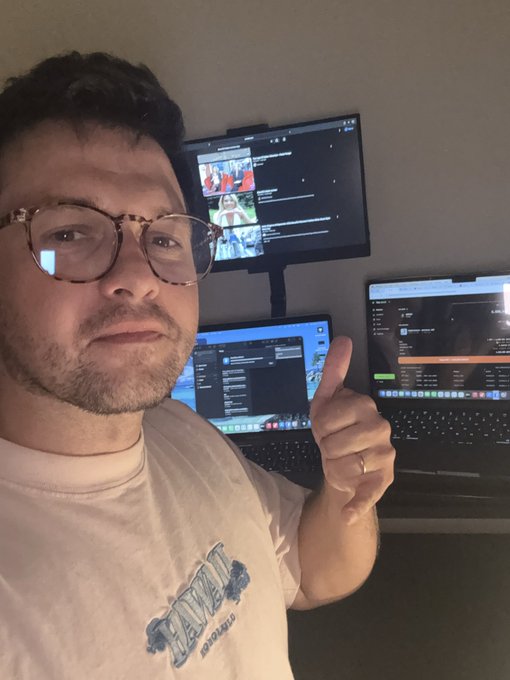 A man wearing glasses and a white t-shirt gives a thumbs-up while sitting in front of a multi-monitor trading setup displaying charts and trading interfaces, celebrating the progress of StonkBrokers as it hits the 1 month and 27 day milestone.