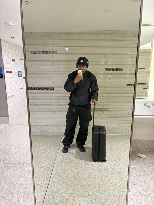 Mirror selfie of a person wearing a black jacket, black pants, black sneakers, and a black NY Yankees cap, holding a phone while standing next to a black rolling suitcase in a tiled public restroom.