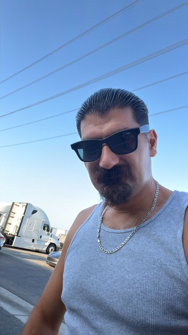 A man with slicked-back dark hair, a goatee, dark sunglasses, and a silver chain necklace wears a light blue tank top, taking a selfie outdoors with power lines and a white semi-truck visible behind him.