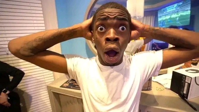 A young man in a white t-shirt makes an exaggerated shocked expression with his mouth open and hands on his head, standing in a room with a TV monitor in the background.