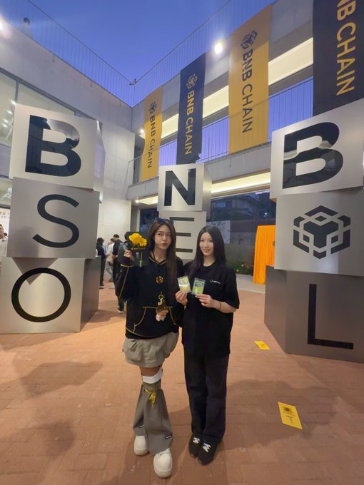 Two young women posing indoors in front of BNB Chain branded signage and a BNB SOLO installation, with one holding a yellow flower and the other holding a green item.