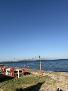 A seaside view in Athens, Greece, featuring red chairs and tables arranged along a rocky shoreline with the calm sea and distant mountains under a clear blue sky. The image complements the post's reference to Vechain ($VET, $VTHO), evoking a relaxing Mediterranean atmosphere likely tied to travel or lifestyle content involving the crypto community.