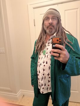 A mirror selfie of a person with long dreadlocks, glasses, and a beanie, wearing a green checkered robe over a printed shirt.