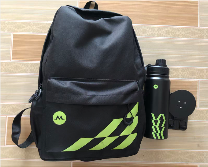 Black backpack and matching water bottle featuring green geometric designs and a stylized 'M' logo, displayed on a tiled floor alongside what appears to be a disc case.
