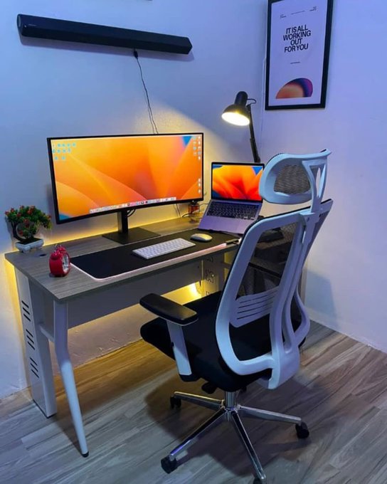A modern home office setup featuring a wooden desk with an ultrawide monitor and laptop both displaying orange wallpapers, accompanied by a white ergonomic chair, a desk lamp, and a framed poster reading 'IT IS ALL WORKING OUT FOR YOU'.