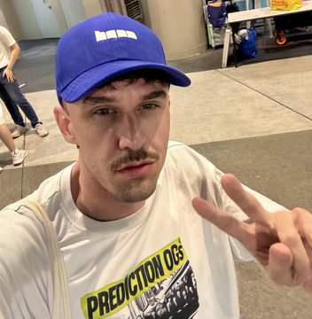 A man in a blue cap and white graphic T-shirt takes a selfie while holding up a two-finger peace sign on a sidewalk.