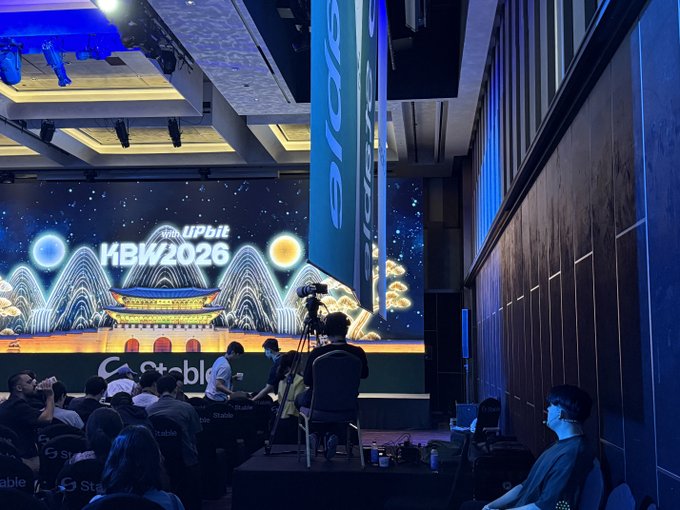 A photograph of a conference event with a large digital backdrop displaying "Upbit KBW2026" alongside an illustration of a traditional Korean building; audience members are seated facing the stage.