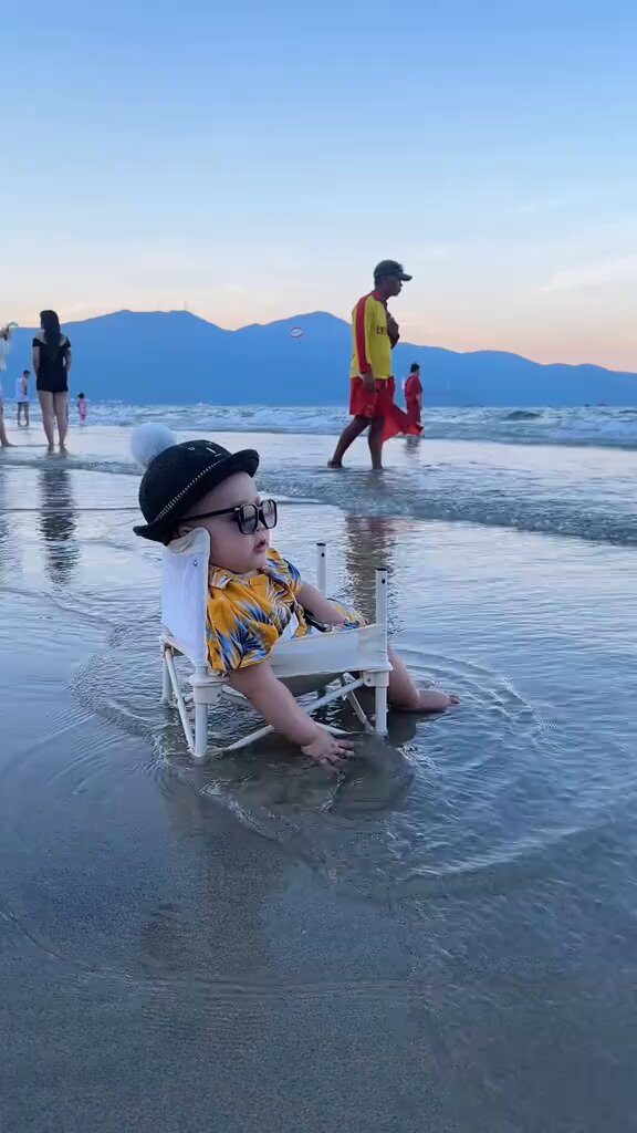 The image features a baby sitting in a white lawn chair at the beach, wearing glasses and a yellow shirt. The background shows people walking along the shoreline near mountains, creating an overall beachy atmosphere. The photo has been shared on social media with the caption "Gm Good morning Fam! Đầu tuần năng lượng nha anh em tiêp tục build mạnh nè !".