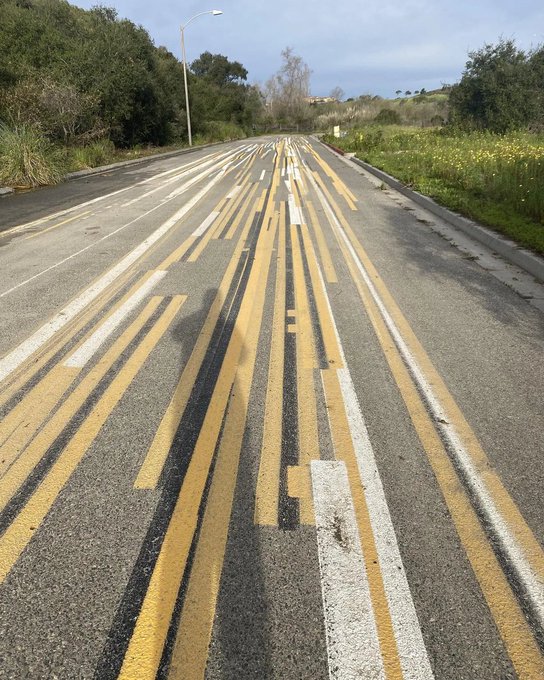 A photograph of an asphalt road with yellow and white lane markings that appear distorted, blurred, or glitched in a streaky pattern down the center, surrounded by greenery and a street lamp under a cloudy sky.