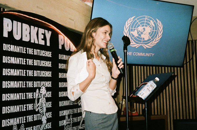 A young woman with long brown hair speaks into a microphone on stage, smiling, with a backdrop reading "PUBKEY - DISMANTLE THE BITLICENSE" and a UN emblem displaying "THE COMMUNITY." The setting suggests a cryptocurrency or blockchain-related event, aligning with the "$dasha" reference to the speaker's identity.
