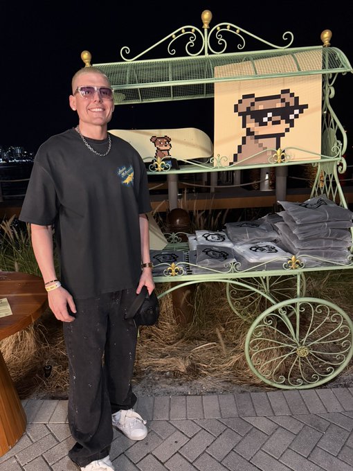 A smiling young man wearing glasses and a black t-shirt with a "Doginal Dogs" logo poses beside an ornate green cart stacked with Doginal Dogs merchandise and featuring a pixel-art dog portrait, celebrating the community's top ranking on X.