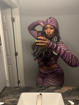 A woman takes a mirror selfie wearing a purple and black zebra-print hooded crop top and matching pants, holding a smartphone in a bathroom setting with a white door and sink visible.