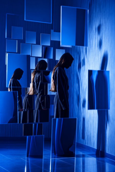 A moody blue-toned photograph of silhouetted figures standing among rectangular mirrored panels that reflect their forms.