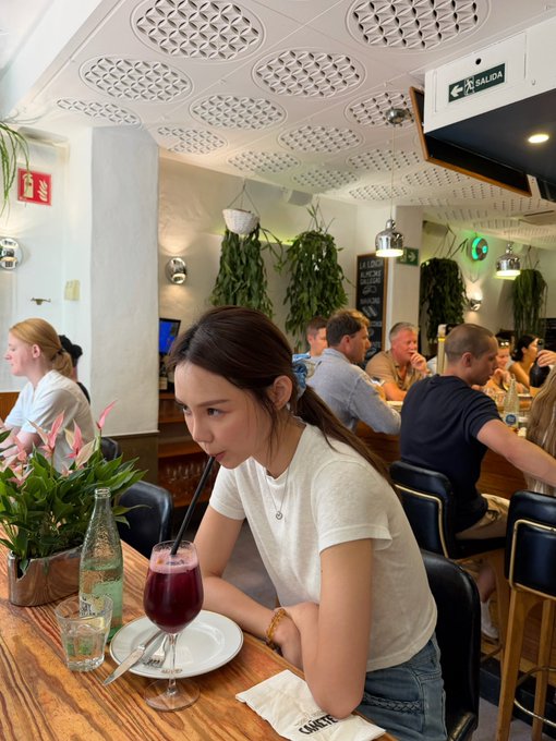 A young woman with dark hair in a white t-shirt sipping a dark red beverage through a straw at a cafe or restaurant table, with other diners, hanging plants, and a 'Salida' emergency exit sign visible in the background.