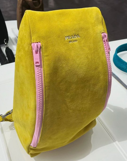A yellow suede Prada handbag featuring two prominent pink zippers is displayed on a white surface, with the "PRADA Milano" logo visible on the front.