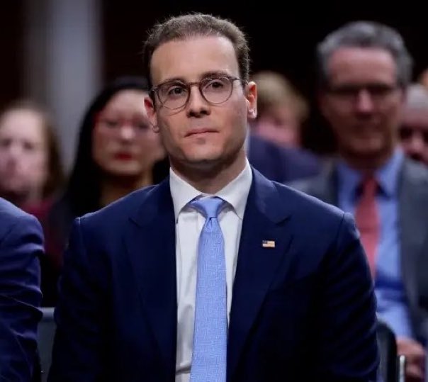 A man in a navy blue suit with a light blue tie and glasses sits in a crowd, with a small American flag pin on his lapel.