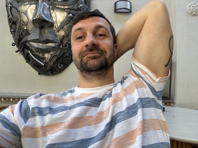 A man with a short beard, wearing a striped t-shirt, reclines with his arm behind his head in front of an industrial-style metallic face wall sculpture.