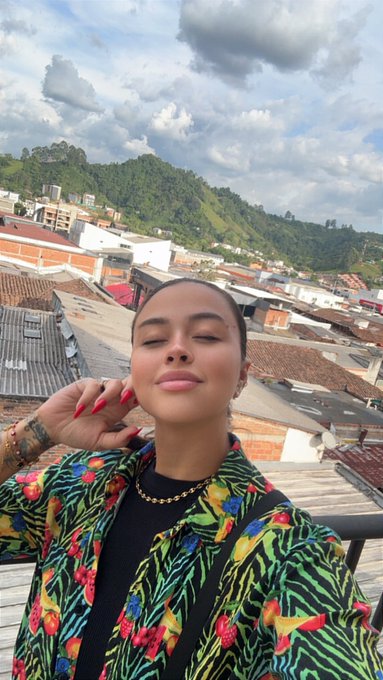 A selfie of a woman with long nails, gold chain, and colorful patterned jacket posing on a rooftop with mountains and a town in the background.