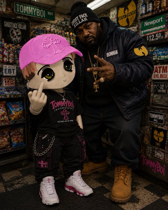 A man wearing a 'GHOSTFACE KILLAH' beanie and 'IRONMAN' jacket poses with a cartoon-style mascot wearing a pink TommyBoy cap, branded apparel, and chains in a store setting decorated with Wu-Tang Clan memorabilia.